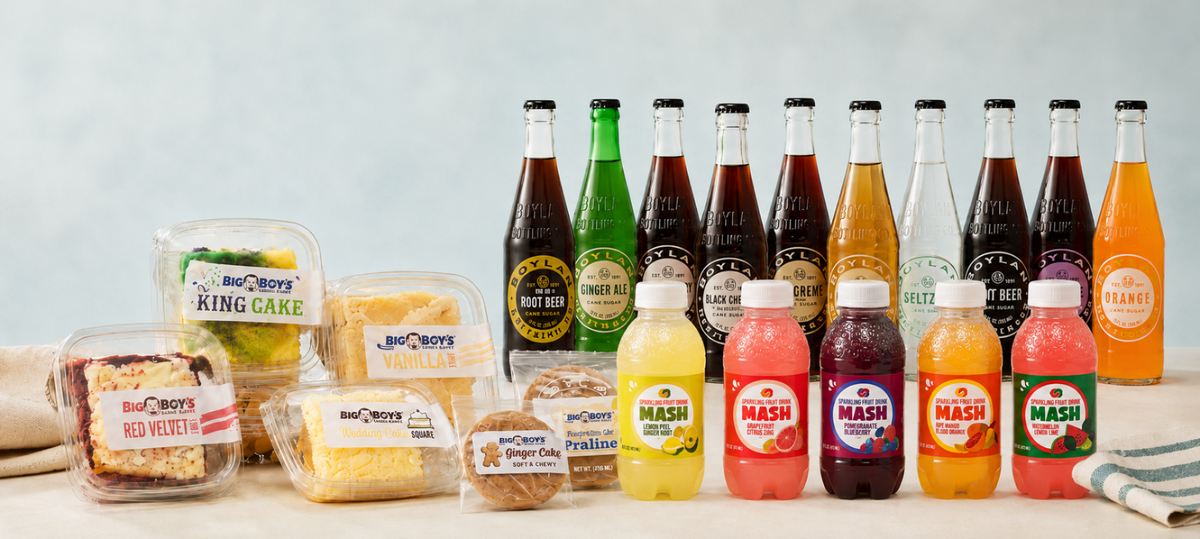 A collection of food products from Big Cheddar's, including packaged cakes, cheeses, and bottled sodas and juices, arranged on a light-colored surface against a pale blue background.
